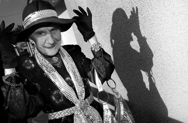 czarnobiałe zdjęcie uśmiechniętej starszej kobiety, która rękoma w rękawiczkach przytrzymuje rondo kapelusza