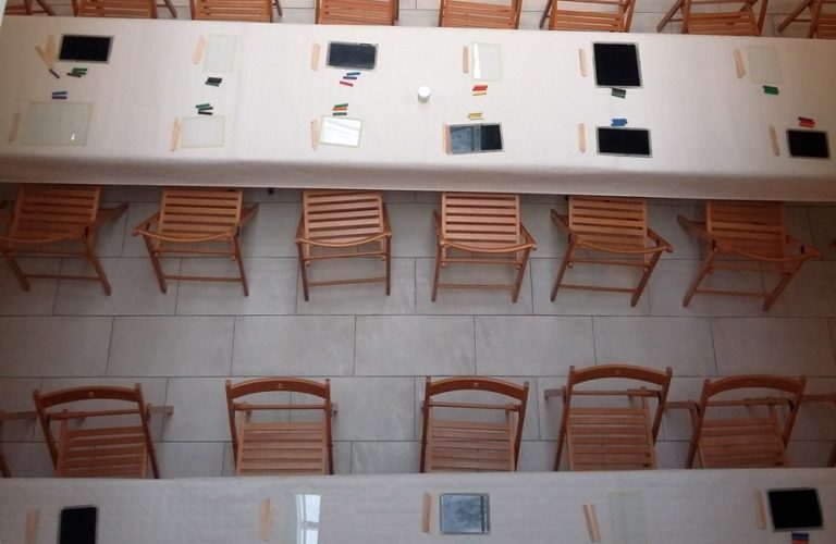 wnętrze galerii - widok z góry na dwa rzędy stolików z ustawionymi przy nich drewnianymi krzesełkami, przygotowanymi na warsztaty plastyczne