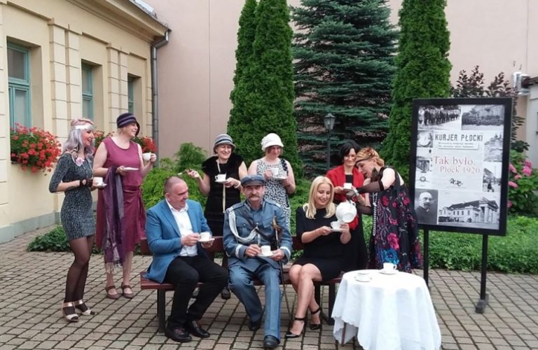 dziedziniec, pozowane zdjęcie - kilka kobiet w strojach stylizowanych stoi, a przed nimi na ławeczce sie Alicja Wasilewska, Michał Kacprzak i mężczyzna przebrany za marszałka Piłsudskieho