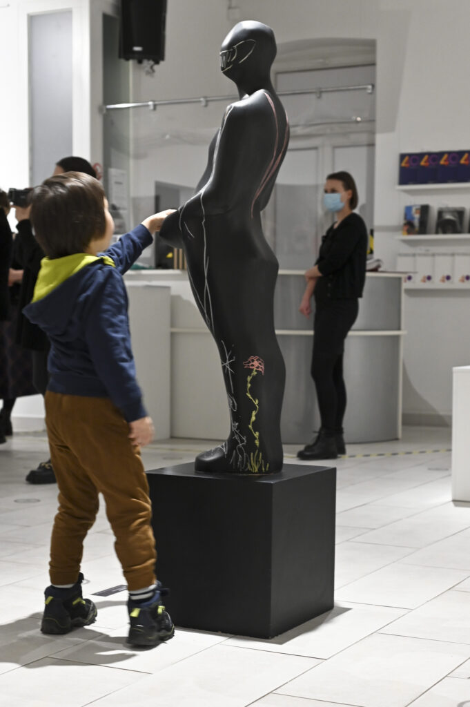wnętrze galerii, kilkuletni chłopiec z ciekawością ogląda czarną rzeźbę, będącą sylwetką człowieka