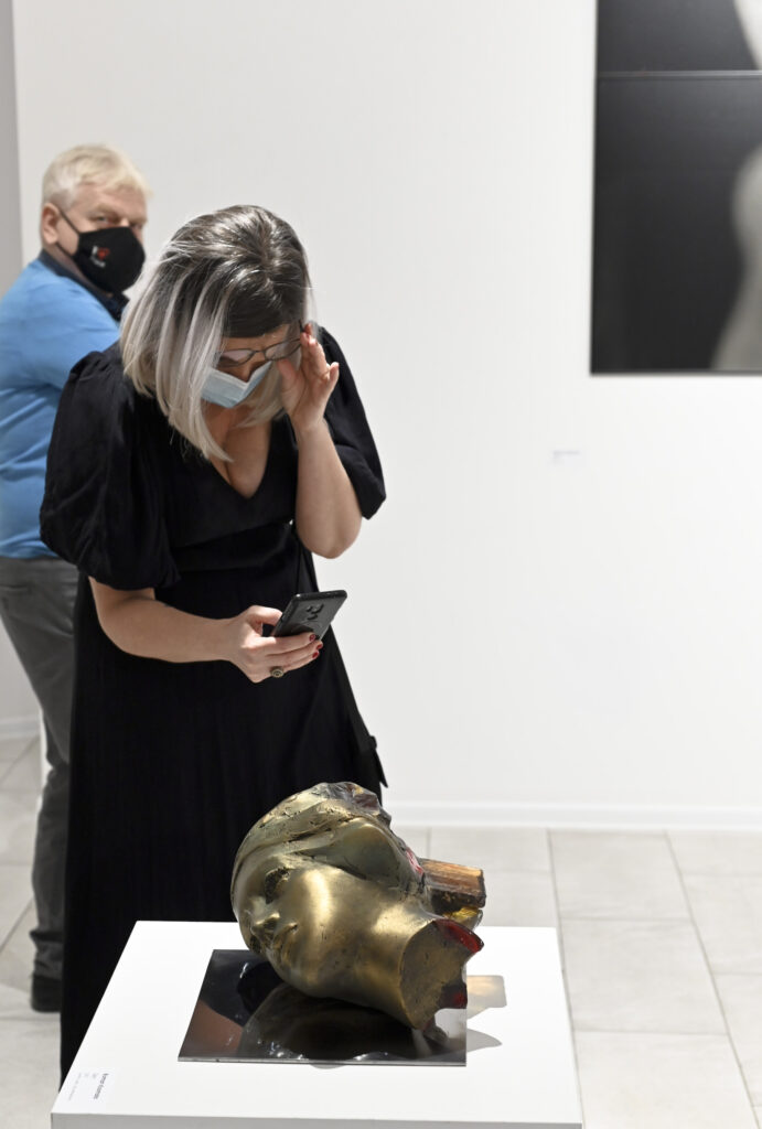 wnętrze galerii, kobieta z uwagą ogląda i fotografuje telefonem komórkowym rzeźbę - to złota, naturalnej wielkości leżąca na boku głowa kobiety