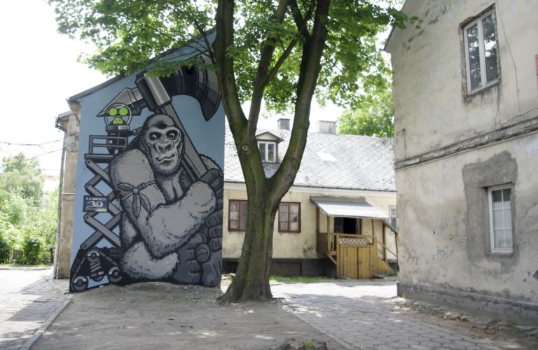 ściana budynku, a na niej mural. Na bladoniebieskim tle w odcieniach szarych popiersie goryla z toporem z gorylem