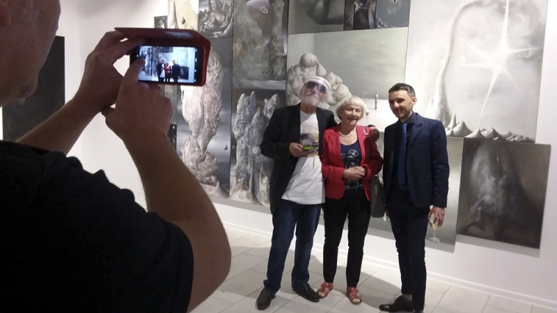 wnętrze galerii, kobieta i mężczyzna pozują z Łukaszem Huculakiem na tle jego obrazów