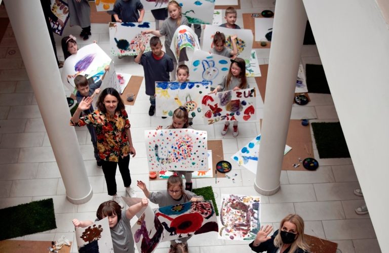Wnętrze Galerii. Zdjęcie zrobione z góry pokazuje grupę dzieci, prezentujących wykonane podczas warsztatów prace