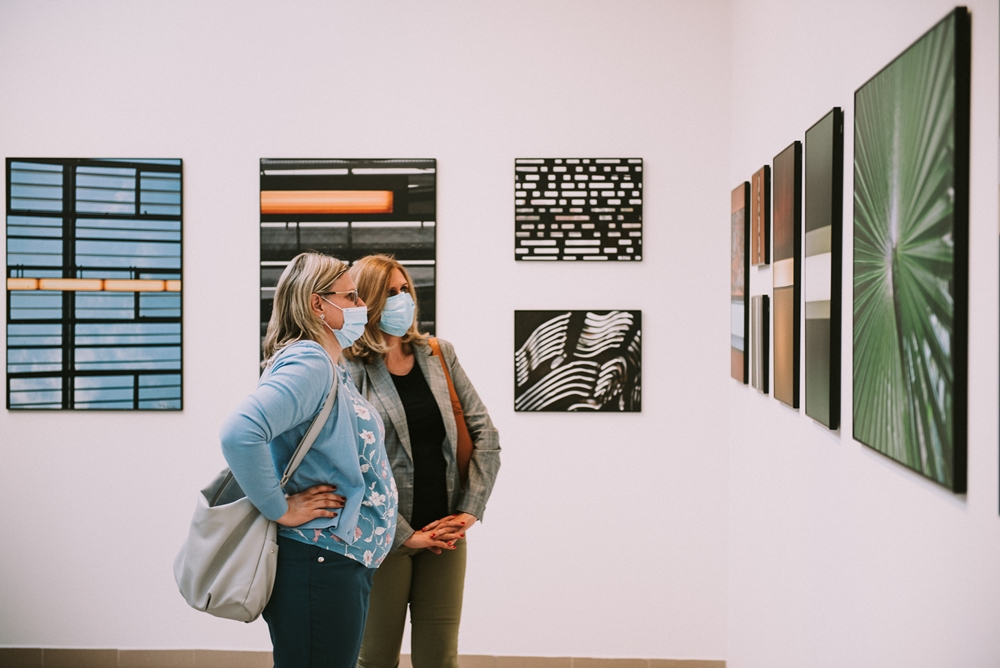 wnetrze galerii, dwie kobiety w maseczkach na twarzach oglądają prezentowane otografie 