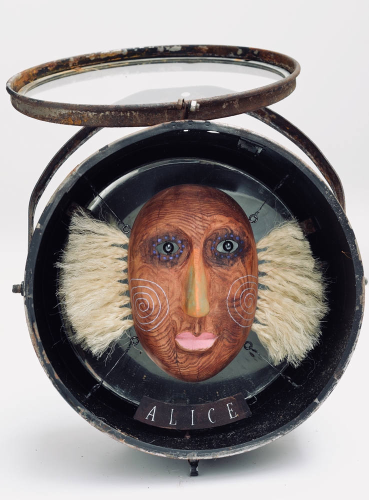 Jüri Mildeberg-Alicja, w przypominającym zegarek-dewizka pudełku namalowana na okrągłym drewienku twarz, po bokach włosy zrobione ze szczeciny