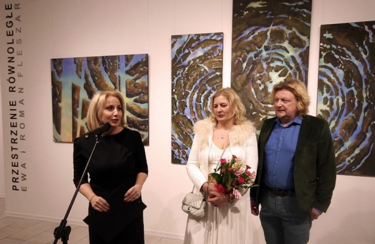 wnętrze galerii na tle obrazów stoją od lewej: przy mikrofonie dyrektor PGS Alicja Wasilewska, obok Ewa i Roman Fleszarowie