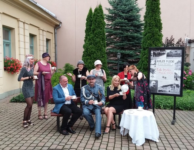 dziedziniec, pozowane zdjęcie - kilka kobiet w strojach stylizowanych stoi, a przed nimi na ławeczce sie Alicja Wasilewska, Michał Kacprzak i mężczyzna przebrany za marszałka Piłsudskieho 