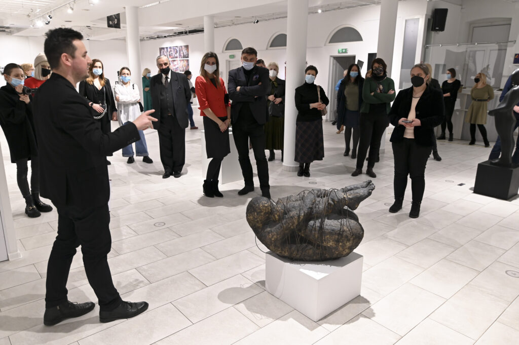 wnętrze galerii, ponad 20 osób słucha Tomasza Jędrzejewskiego, który opowiada o kamiennej rzeźbie skulonego człowieka, obwiązanego drutem