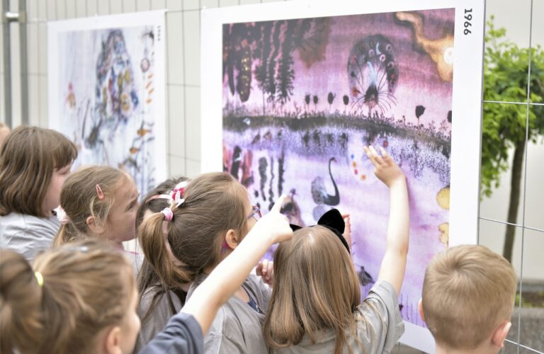 Dziedziniec Galerii. Grupa dzieci ogląda wystawę plenerową i obrazek, na którym są kolorowe pawie