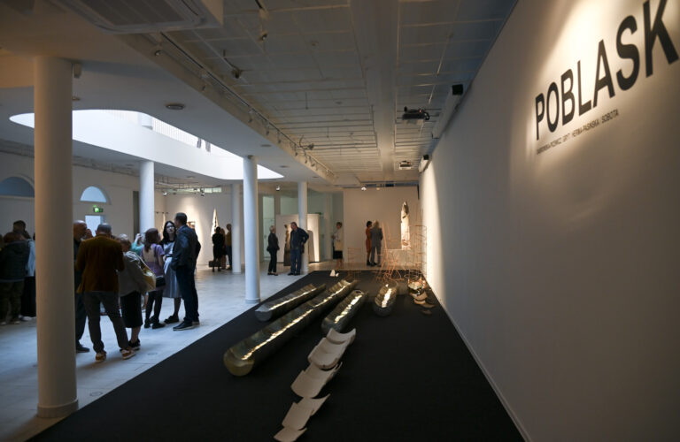 wnętrze galerii, na zdjęciu instalacja ceramiczna przedstawiająca zwalone pnie drzew, nad nimi na ścianie tytuł wystawy - poblask