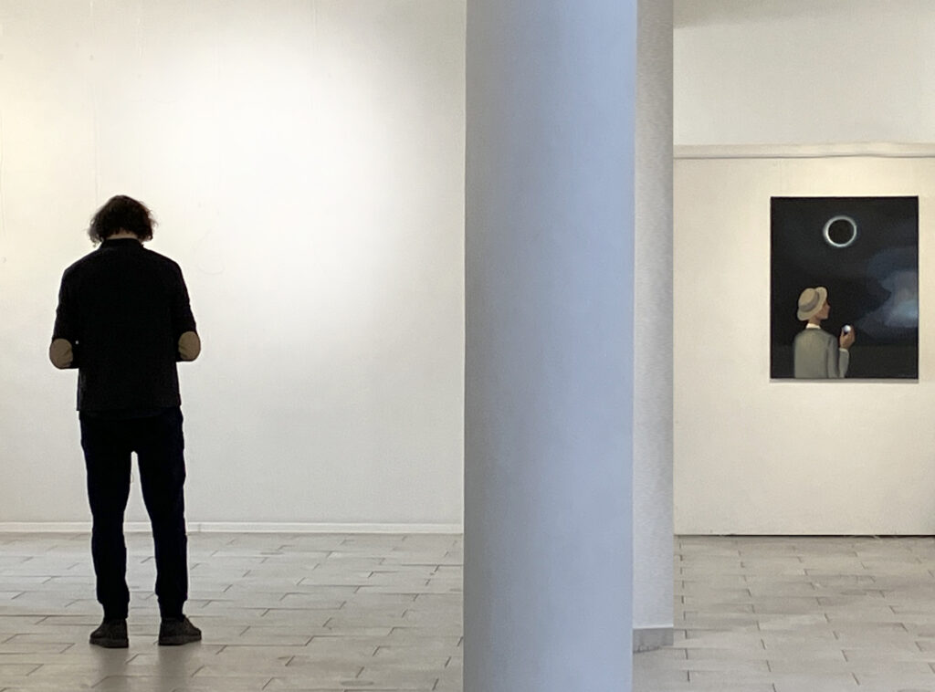 wnętrze galerii z lewej strony na tle pustej ściany stoi tyłem mężczyzna, pośrodku kolumna, a z prawej strony na ścianie wisi duży obraz